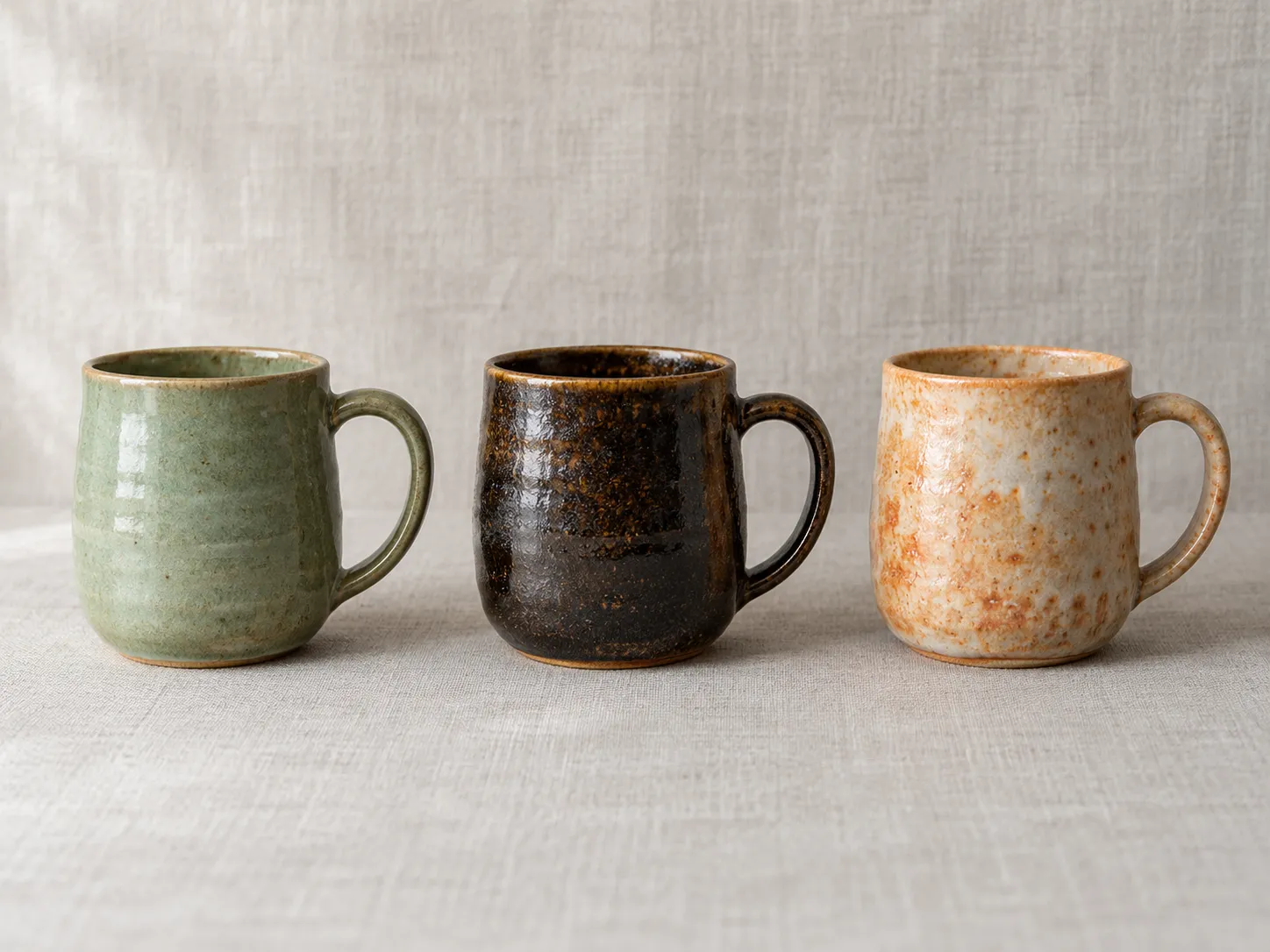 Three handmade mugs in celadon, tenmoku and shino.